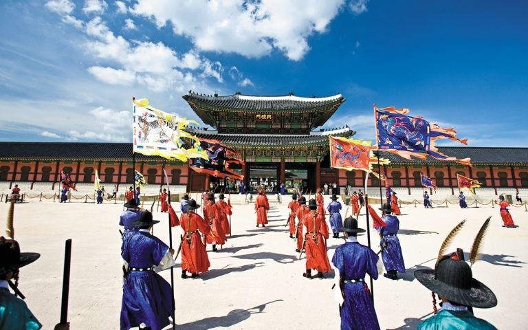 Traditional Korean palace with a ceremonial procession under a blue sky with clouds.