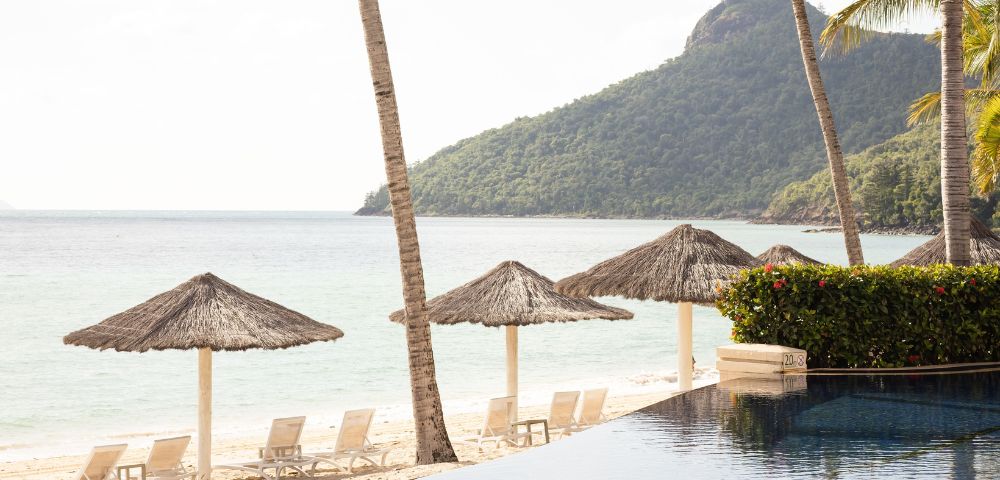 Tropical beach resort with pool, straw umbrellas, palm trees, and a mountain backdrop at the Beach Club Hamilton Island My Queensland.