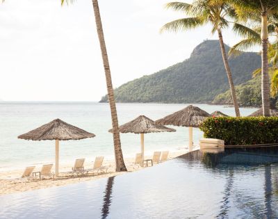 Tropical beach resort with pool, straw umbrellas, palm trees, and a mountain backdrop at the Beach Club Hamilton Island My Queensland.
