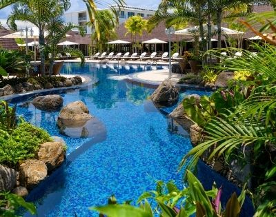 Tropical resort pool with loungers, surrounded by lush plants and palm trees.