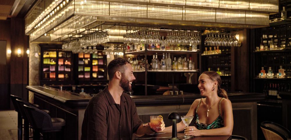 Two individuals sitting at a bar with shelves of bottles and hanging glasses.