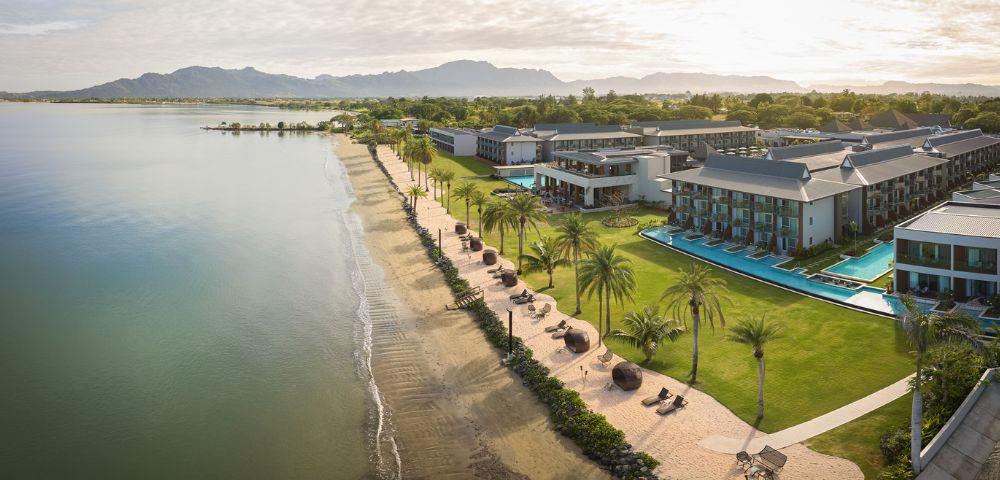 Aerial view of a beachside resort with palm trees, pool, and mountains in the background.