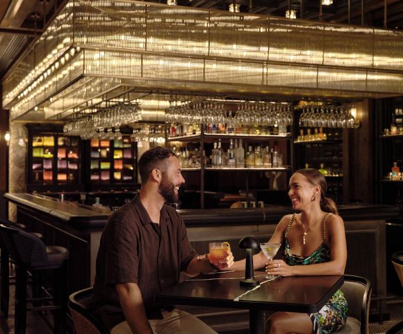 Two individuals sitting at a bar with shelves of bottles and hanging glasses.