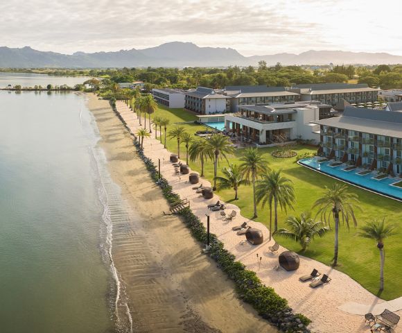 Aerial view of a beachside resort with palm trees, pool, and mountains in the background.