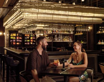 Two individuals sitting at a bar with shelves of bottles and hanging glasses.