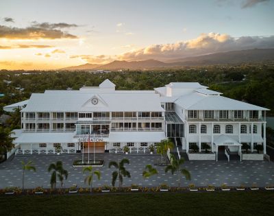 Aerial view of a large white hotel building at sunset with mountains in the background. Sheraton Samoa Aggie Grey’s Hotel and Bungalows.