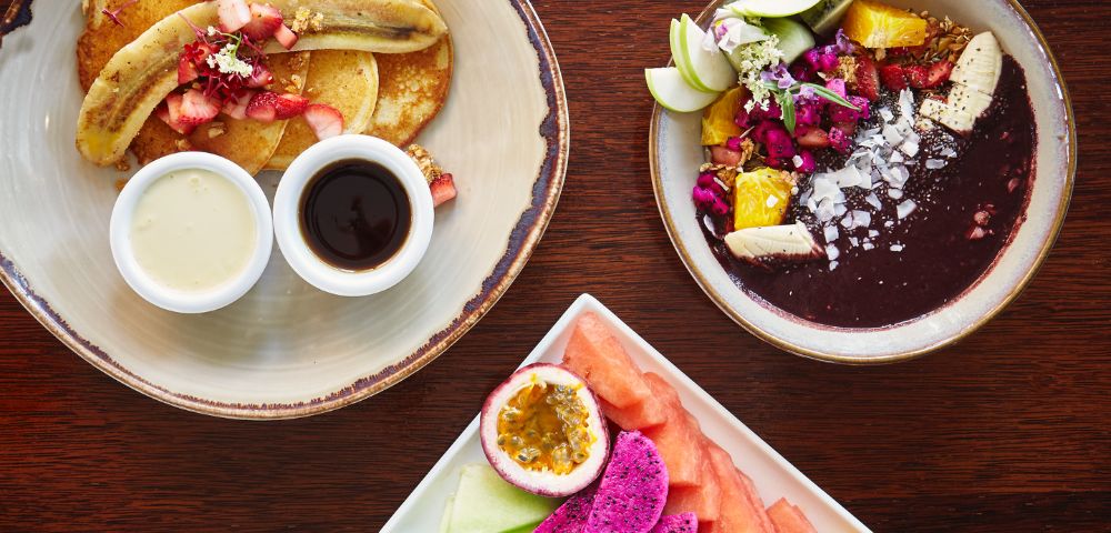 Assorted breakfast plates with pancakes, fruit, and acai bowl.