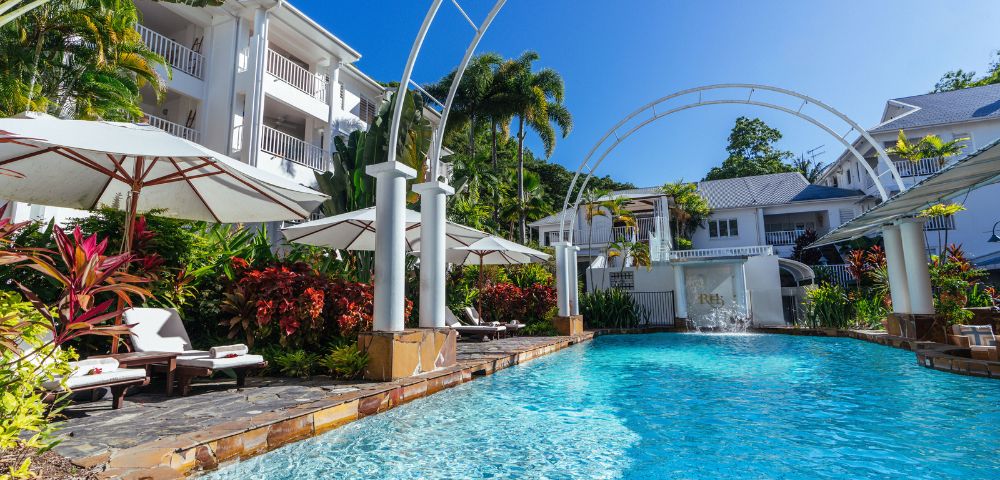 Luxurious resort swimming pool with loungers, surrounded by tropical plants and white buildings.