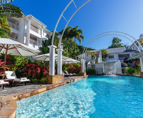 Luxurious resort swimming pool with loungers, surrounded by tropical plants and white buildings.