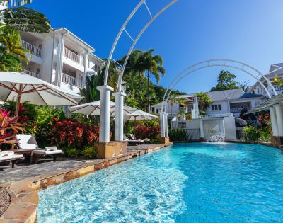 Luxurious resort swimming pool with loungers, surrounded by tropical plants and white buildings.