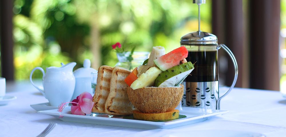 A breakfast tray with toast, fresh fruit in a coconut bowl, a French press, and a milk jug.