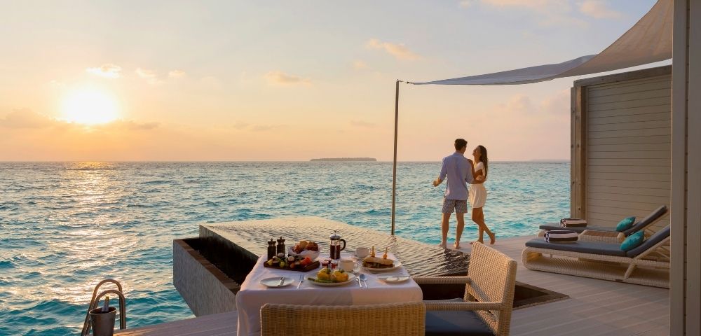 A couple enjoying a sunset by an infinity pool with a dinner table set nearby.