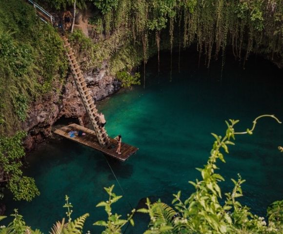 Wooden platform with a ladder over a serene blue water hole, surrounded by lush greenery.