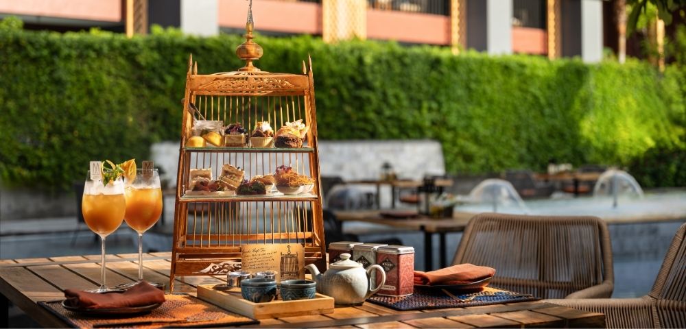 Outdoor table with a birdcage-shaped dessert stand, two cocktails, and hotel backdrop.