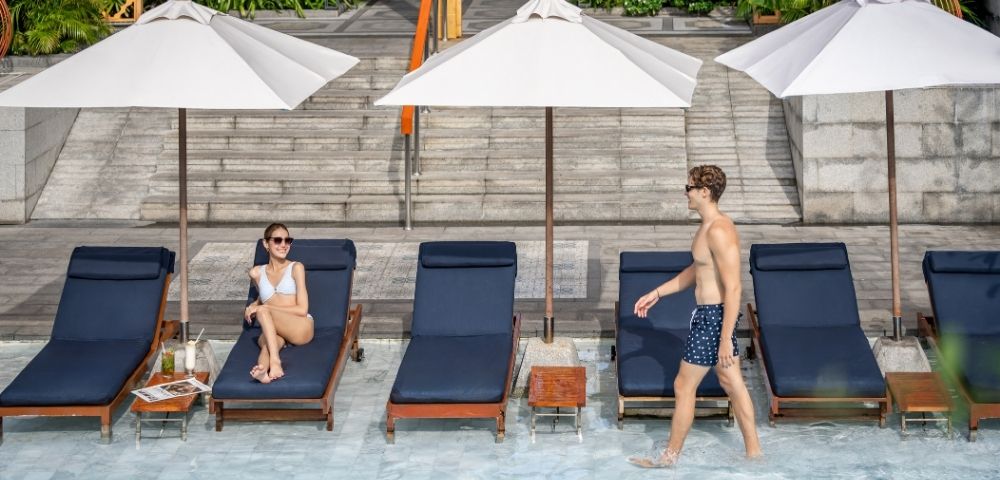 A woman lounging by a pool and a man walking by, with sun umbrellas and deck chairs.