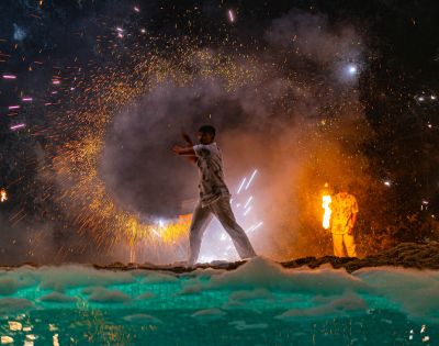 Man with a sparkler amidst fireworks and smoke at night, another person in the background.