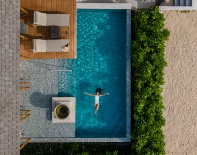Top-down view of a person floating in a pool next to a sandy area with chairs and plants.