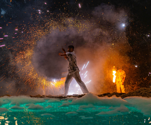 Man with a sparkler amidst fireworks and smoke at night, another person in the background.