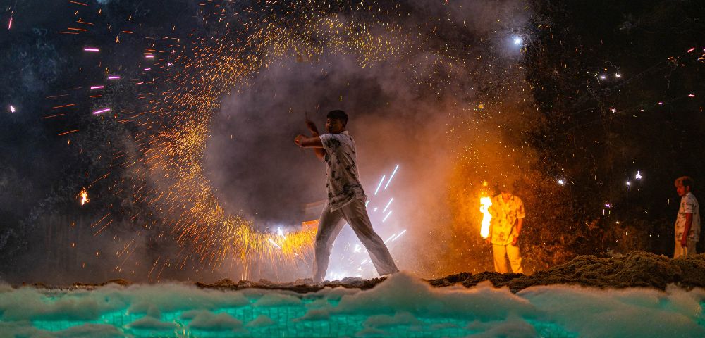 Man with a sparkler amidst fireworks and smoke at night, another person in the background.
