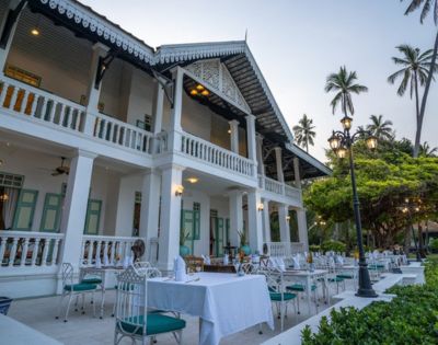 Outdoor dining setup at a colonial-style building with palm trees and string lights at twilight.