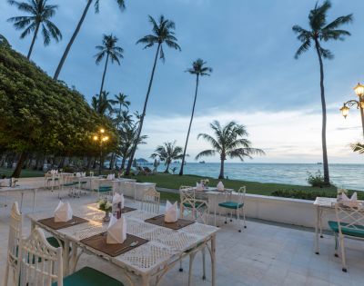 Outdoor beachfront dining area with tables set up under tall palm trees during dusk.