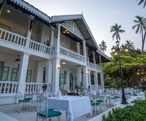 Outdoor dining setup at a colonial-style building with palm trees and string lights at twilight.