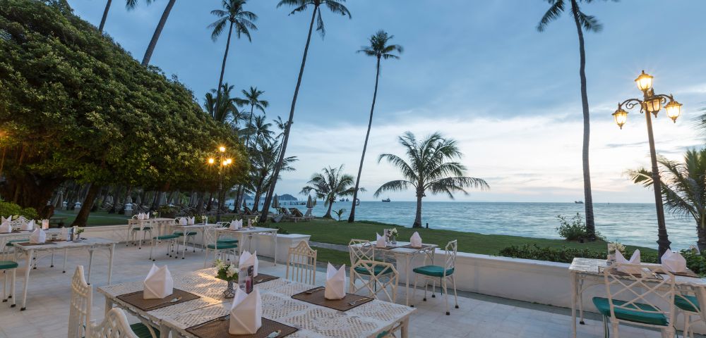 Outdoor beachfront dining area with tables set up under tall palm trees during dusk.