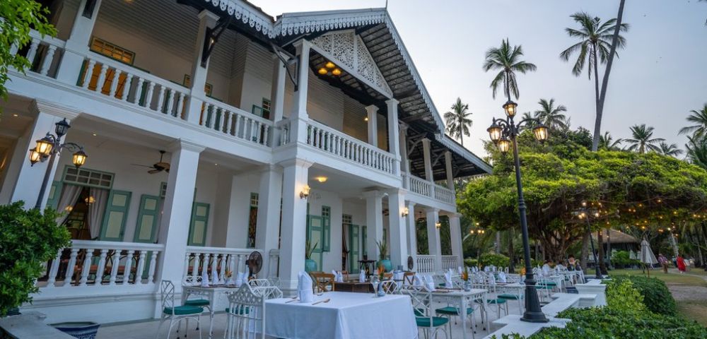 Outdoor dining setup at a colonial-style building with palm trees and string lights at twilight.