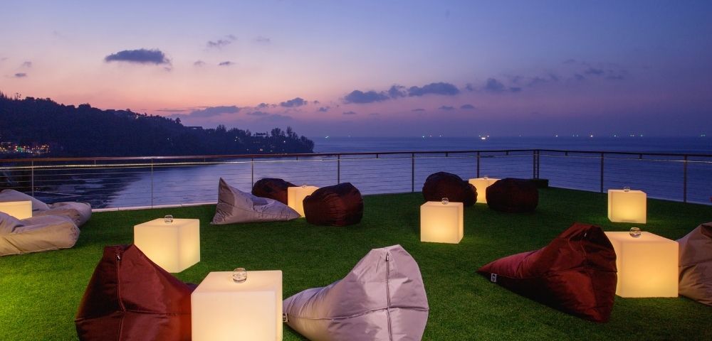 Outdoor lounge with bean bags and lamps on grass overlooking the sea at dusk.