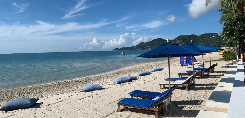 Beach scene with sun loungers and umbrellas on sandy shore against a backdrop of clear blue sky and hills.