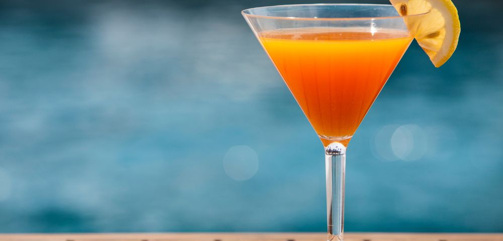 Orange cocktail with lemon garnish in a martini glass on a wooden surface, blurred pool background.