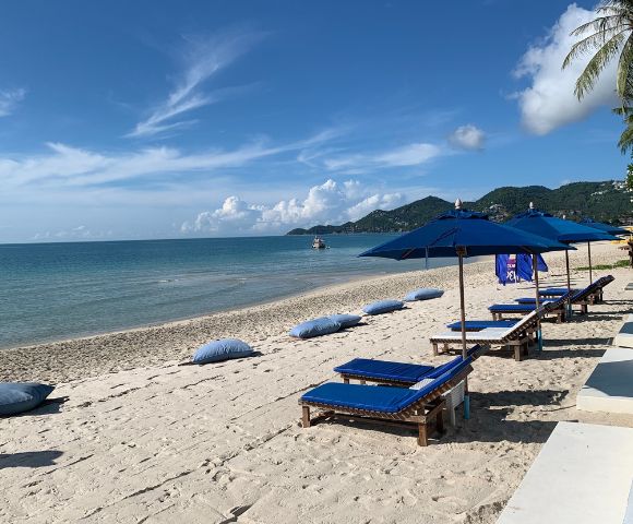 Beach scene with sun loungers and umbrellas on sandy shore against a backdrop of clear blue sky and hills.