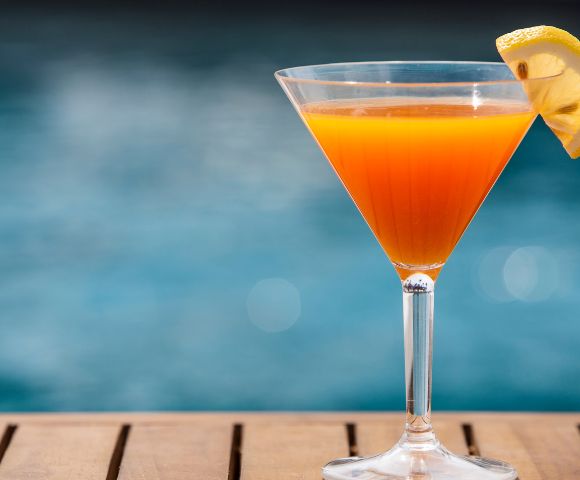 Orange cocktail with lemon garnish in a martini glass on a wooden surface, blurred pool background.