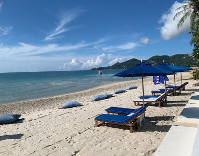 Beach scene with sun loungers and umbrellas on sandy shore against a backdrop of clear blue sky and hills.