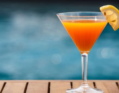 Orange cocktail with lemon garnish in a martini glass on a wooden surface, blurred pool background.