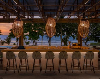 Beachside bar at dusk with wicker pendant lights and a row of stools facing the ocean.