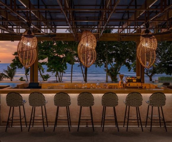 Beachside bar at dusk with wicker pendant lights and a row of stools facing the ocean.
