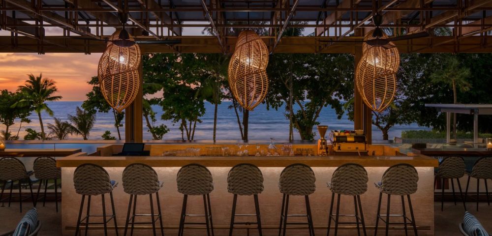 Beachside bar at dusk with wicker pendant lights and a row of stools facing the ocean.