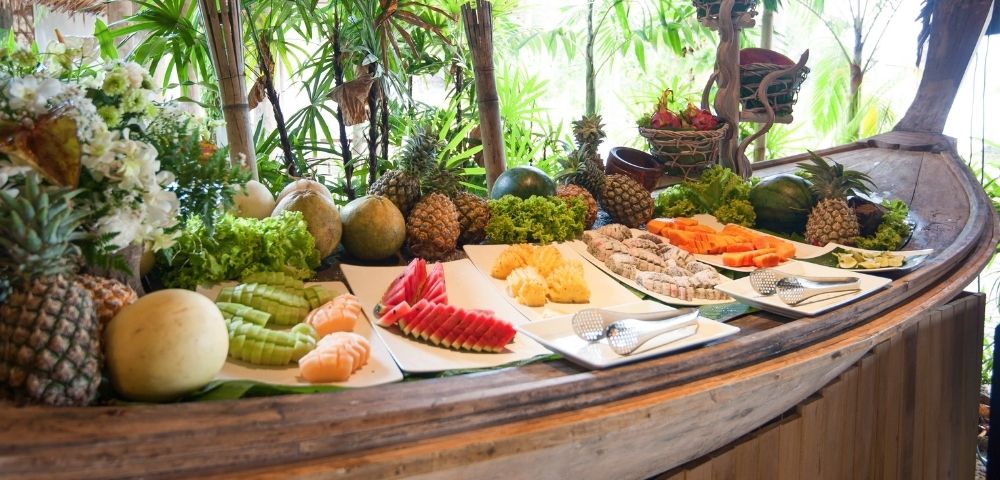 A tropical buffet with an array of fruits under a thatched roof.
