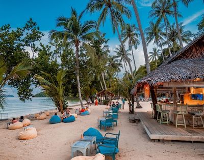 Tropical beach bar with lounge chairs and bean bags on sandy shore lined with palm trees.