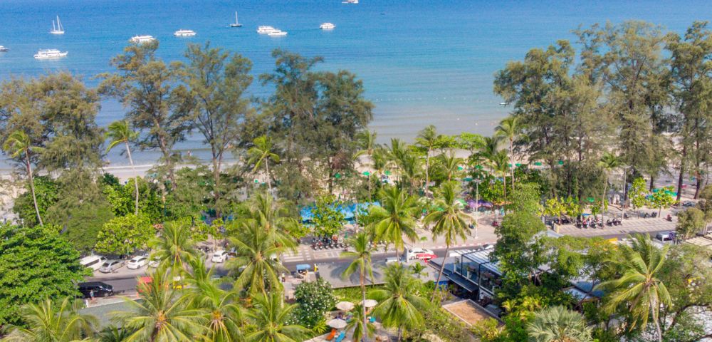 Aerial view of a tropical resort with a pool near a beach lined with palm trees and boats in the sea.