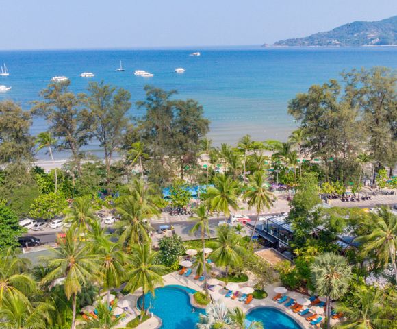 Aerial view of a tropical resort with a pool near a beach lined with palm trees and boats in the sea.