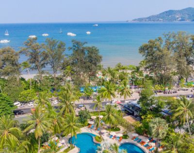 Aerial view of a tropical resort with a pool near a beach lined with palm trees and boats in the sea.