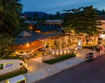 Twilight view of a resort entrance with lit-up café and street, surrounded by tropical trees.