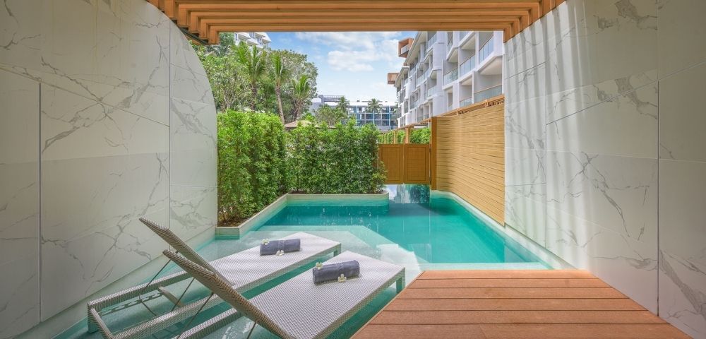 Modern outdoor patio with a pool, wooden deck, loungers, and greenery under a pergola.