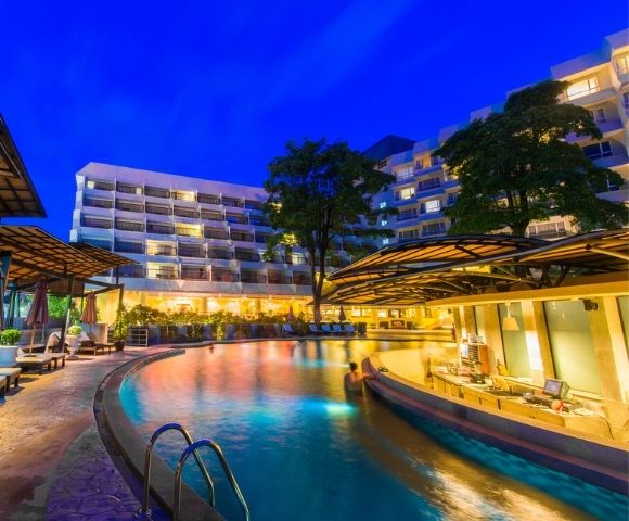 Illuminated resort pool at twilight with buildings in the background.