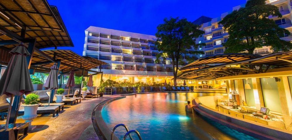 Illuminated resort pool at twilight with buildings in the background.