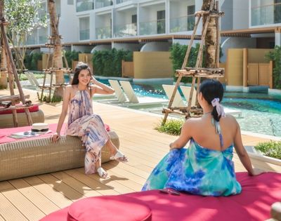Two people in colorful dresses sitting by a resort pool with sun loungers and palm trees.