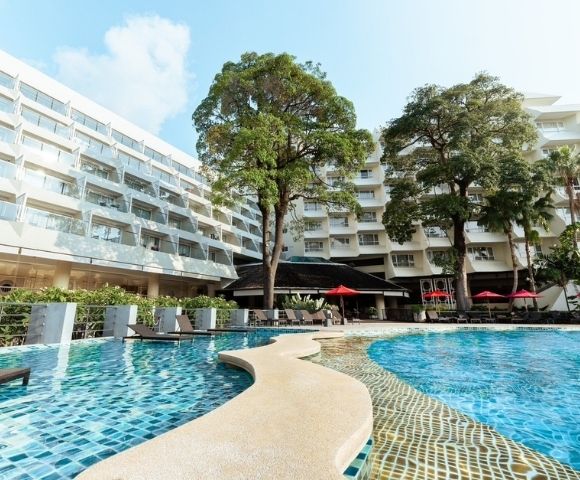 A resort pool with blue mosaic tiles, surrounded by white buildings with balconies and trees.