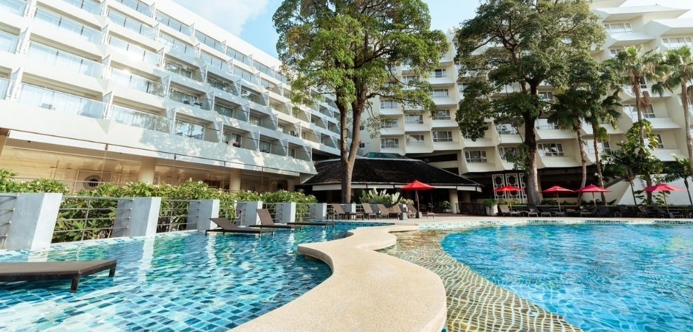 A resort pool with blue mosaic tiles, surrounded by white buildings with balconies and trees.
